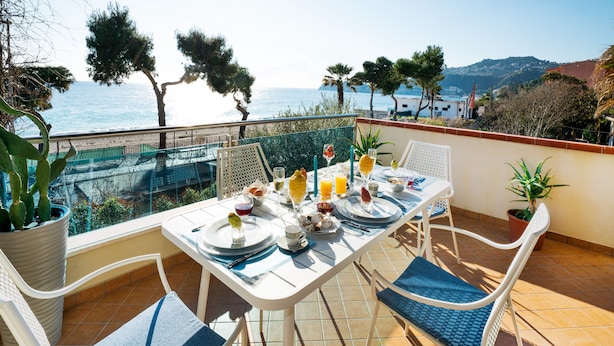 A dining table set on a balcony with a view of the sea and hills.