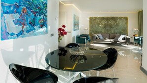 A modern living room with a glass dining table, black chairs, and a sofa with cushions. There is a large abstract painting on the wall and a vase with red flowers on a side table.