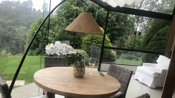 Une table ronde en bois, sur laquelle sont posés un vase en verre et un verre à vin, entourée de chaises en osier avec des coussins, le tout sous une pergola agrémentée d’une lampe suspendue.