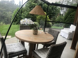 Ett runt träbord med en vas av glas och ett vinglas, omgivet av flätade stolar med kuddar, under en pergola med en hängande lampa.