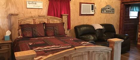 Interior de uma cabana de madeira com uma cama, uma poltrona de couro, uma placa com a inscrição 'Welcome Cabin' e um ar condicionado fixado na parede.