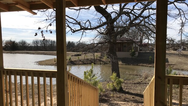 En träveranda med utsikt över en flod, med ett träd och ett hus i bakgrunden.