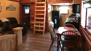 En stuga med trägolv, ett matbord med stolar, ett köksområde med kylskåp och spis, en trätrappa och en soffa klädd i läder.