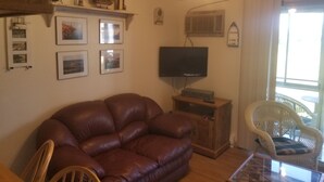 A living room with a brown leather sofa, a television on a wooden stand, a wicker chair, and a wall decorated with framed pictures and a ship model. - Great place for fisherman and bird watchers (Oak Harbor)
