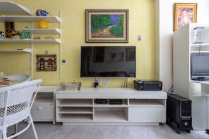 A room with a TV on a white stand, a bookshelf with various items, a wicker chair, and a small table with a vase and a book. - Ipanema 2 bedrooms lagoon and beach (Ipanema)