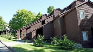 Une maison en bois à deux étages, de couleur brune, avec un toit en pente, un porche à l’avant et un petit jardin verdoyant.