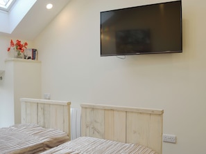 A bedroom with a TV mounted on the wall, wooden headboards, a shelf with books and flowers, and a skylight.