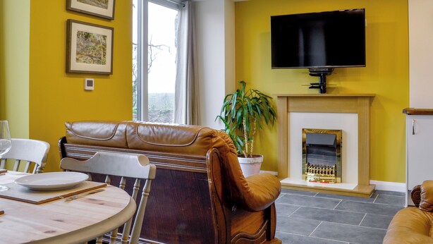 A living room with a sofa, a dining table with chairs, a fireplace, a television mounted on the wall, and framed pictures on the wall. - Dutch Barn One - UK12872 (Ymwlch, near Porthmadog)