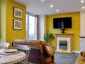 Un salon comprenant un canapé, une table à manger avec des chaises, une cheminée, un téléviseur fixé au mur et des tableaux encadrés accrochés au mur.