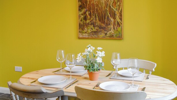 Uma mesa de jantar posta com pratos, copos e talheres, uma planta em vaso e um quadro na parede.
