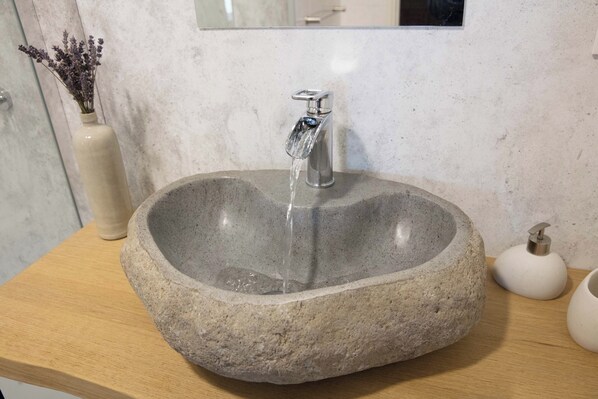Un lavamanos de piedra con agua corriente, una encimera de madera, un jarrón con flores secas y un dispensador de jabón.