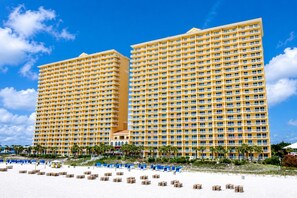 A large hotel with multiple floors and balconies, located near a beach with sun loungers and beach chairs.