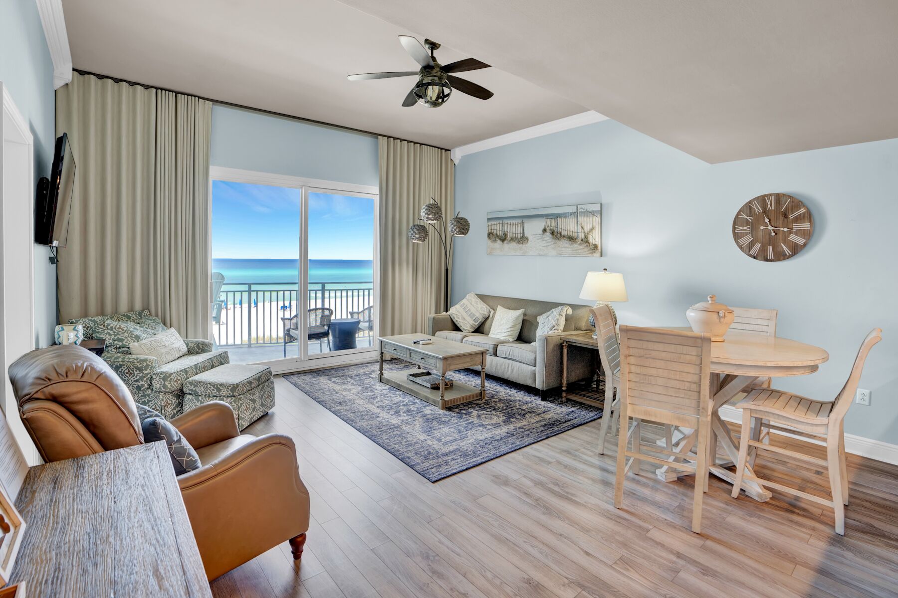 Ein geräumiges Wohnzimmer mit großem Fenster zum Strand, einem Deckenventilator, einem Essbereich mit Tisch und Stühlen sowie einer Sofagarnitur.