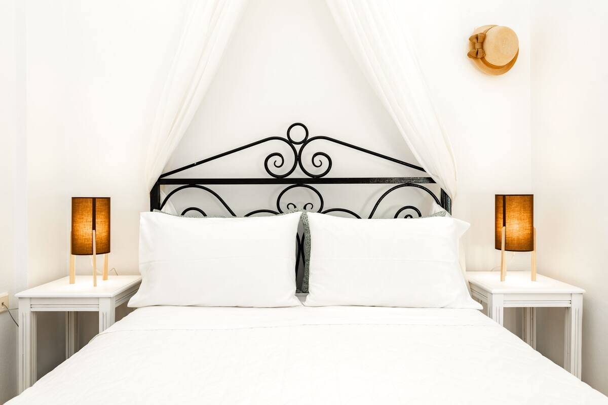 Een netjes opgemaakt bed met witte lakens, een zwart metalen hoofdbord en twee bedlampen op witte nachtkastjes.