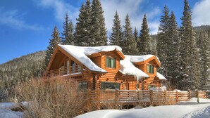 Une cabane en rondins dont le toit est recouvert de neige, entourée de grands pins et surplombée d’un ciel bleu clair.