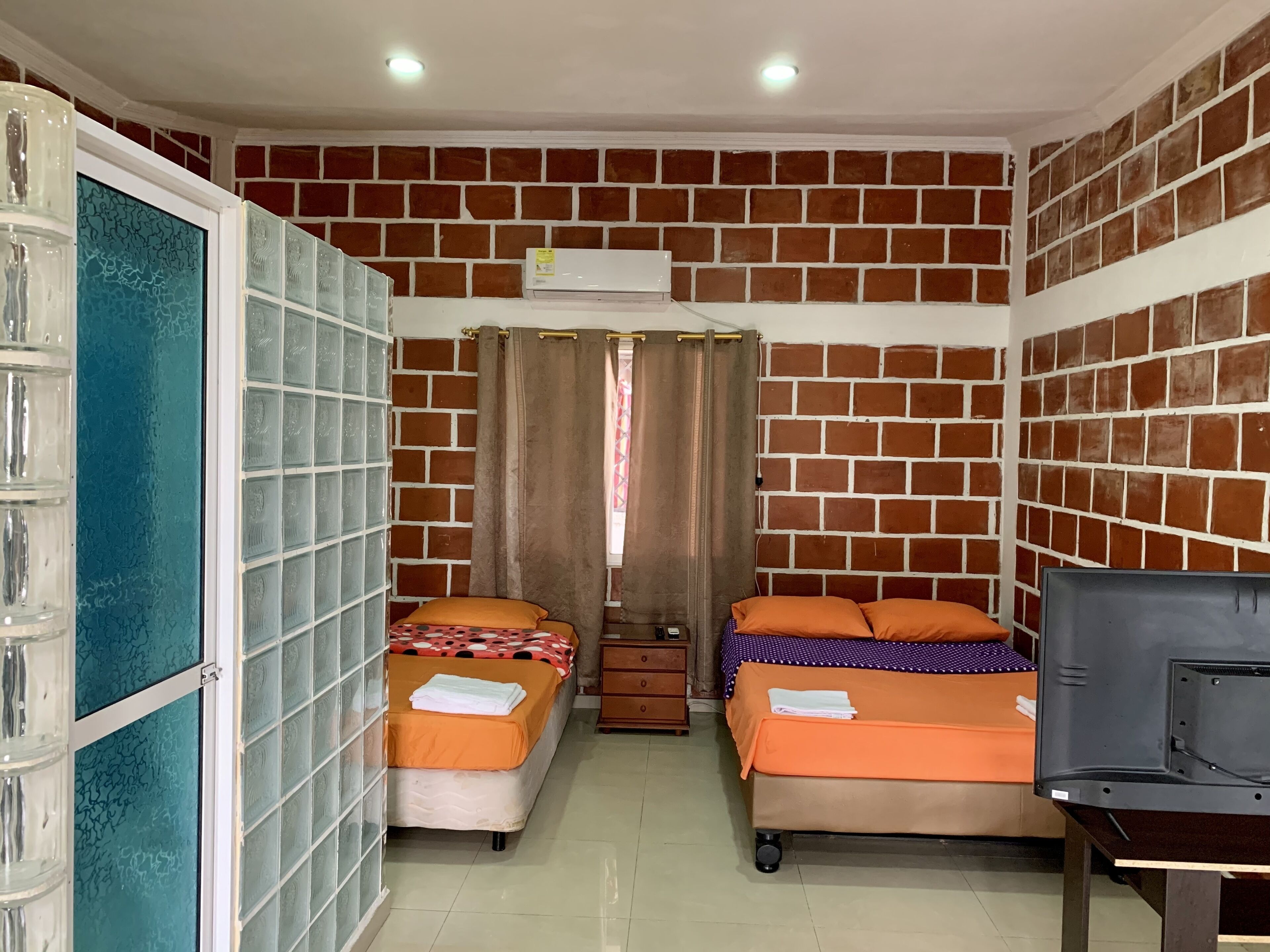 A room with brick-patterned walls, two beds with orange bedding, a TV on a stand, and an air conditioning unit.