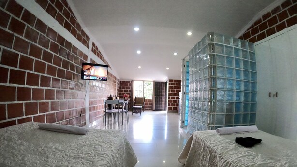 Quarto de hotel com duas camas, televisão fixada na parede, área de refeições com mesa e cadeiras, e uma grande janela com painéis de vidro azulados.