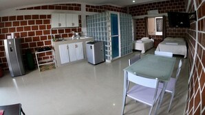 Cozinha compacta com armários brancos, uma geladeira e um micro-ondas. No quarto vizinho, há duas camas com roupa de cama branca.