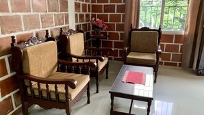 Quarto com parede de tijolos, duas cadeiras de madeira com assentos acolchoados, uma mesa de madeira com tampo de vidro e uma janela com vista para vegetação.