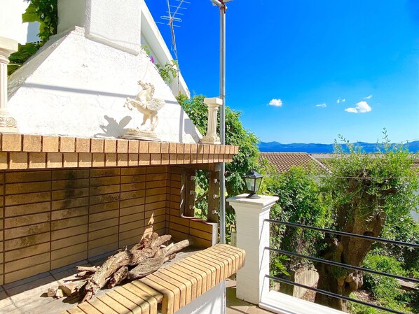 Een balkon met een bakstenen openhaard, een decoratief haanbeeld en uitzicht op zee en bergen.