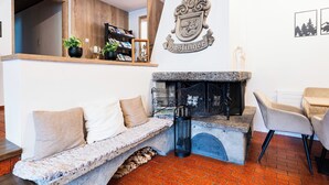 Una sala de estar acogedora con chimenea de piedra, estante de madera con libros y plantas, banco de piedra y pared blanca con un escudo.