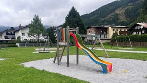 Un parque infantil con un tobogán de colores y estructuras para escalar, rodeado de césped y con montañas al fondo.