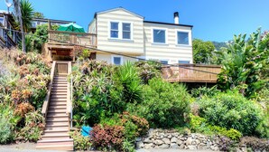 A two-story house with a wooden deck, a stone wall, and a variety of plants. - Sunny Beach Cottage with Amazing Views (Stinson Beach)