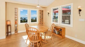 A dining area with a wooden table and chairs, a bookshelf, and a view of the sea through large windows. - Sunny Beach Cottage with Amazing Views (Stinson Beach)