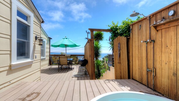 A wooden deck with a hot tub, a patio table with chairs, and a green umbrella. - Sunny Beach Cottage with Amazing Views (Stinson Beach)