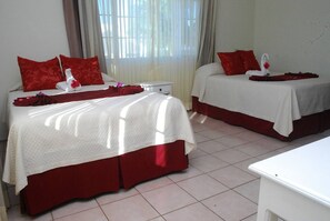 Quarto de hotel com duas camas, cada uma com roupa de cama e almofadas vermelhas, uma mesa de cabeceira branca e uma janela com cortinas.
