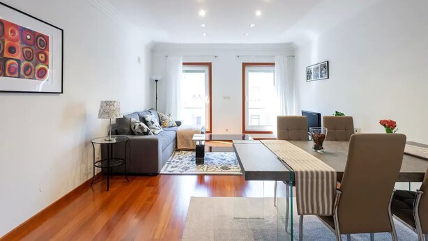 Una sala de estar moderna con sofá, mesa de centro y zona de comedor. Hay dos ventanas con cortinas, una lámpara y un cuadro en la pared.