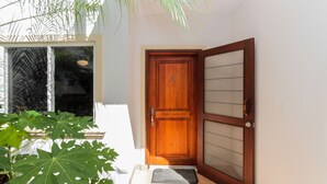 A wooden door with a glass panel, a mat at the entrance, and a large green plant in the foreground. - Ocean Breeze 11 (Sardinal)