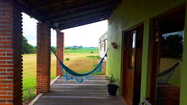 Un porche de madera con una hamaca, una planta en maceta y vistas a un campo verde.