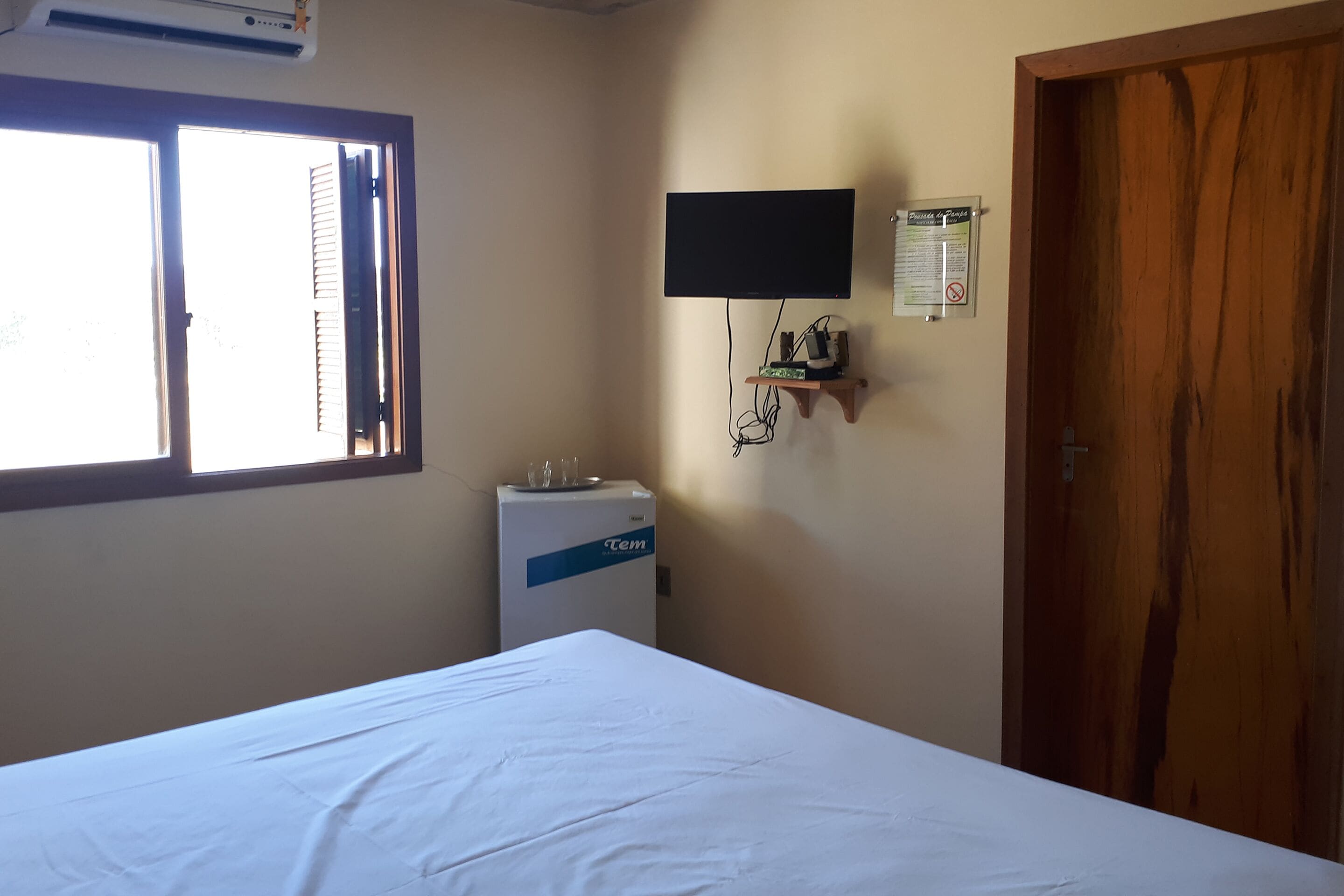 A hotel room with a bed, a TV mounted on the wall, a small shelf with items, and a wooden door.