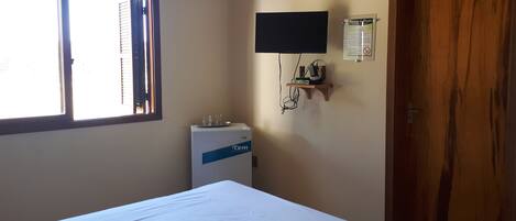 A hotel room with a bed, a TV mounted on the wall, a small shelf with items, and a wooden door.