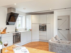 A modern kitchen with white cabinets, a built-in oven, and a dining area with a wooden table and a grey sofa.