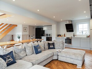 A modern kitchen with a built-in oven, a dining area with a wooden table, and a grey sectional sofa with decorative pillows.