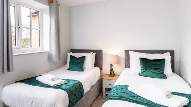 Ein Hotelzimmer mit zwei Betten, jedes mit einer grünen Tagesdecke und weißen Kissen. Es gibt ein Fenster mit Vorhängen, einen Nachttisch mit einer Lampe und eine kleine Statue auf dem Tisch.