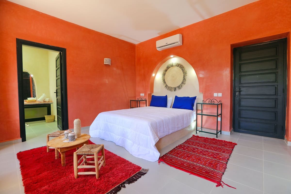 Um quarto com uma cama, uma mesa com cadeiras, um tapete vermelho e uma porta preta.