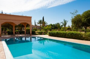 A swimming pool with a clear blue water surface, surrounded by a landscaped garden with various plants and trees, and a building with arches in the background. - Atlas Widan (Marrakech)