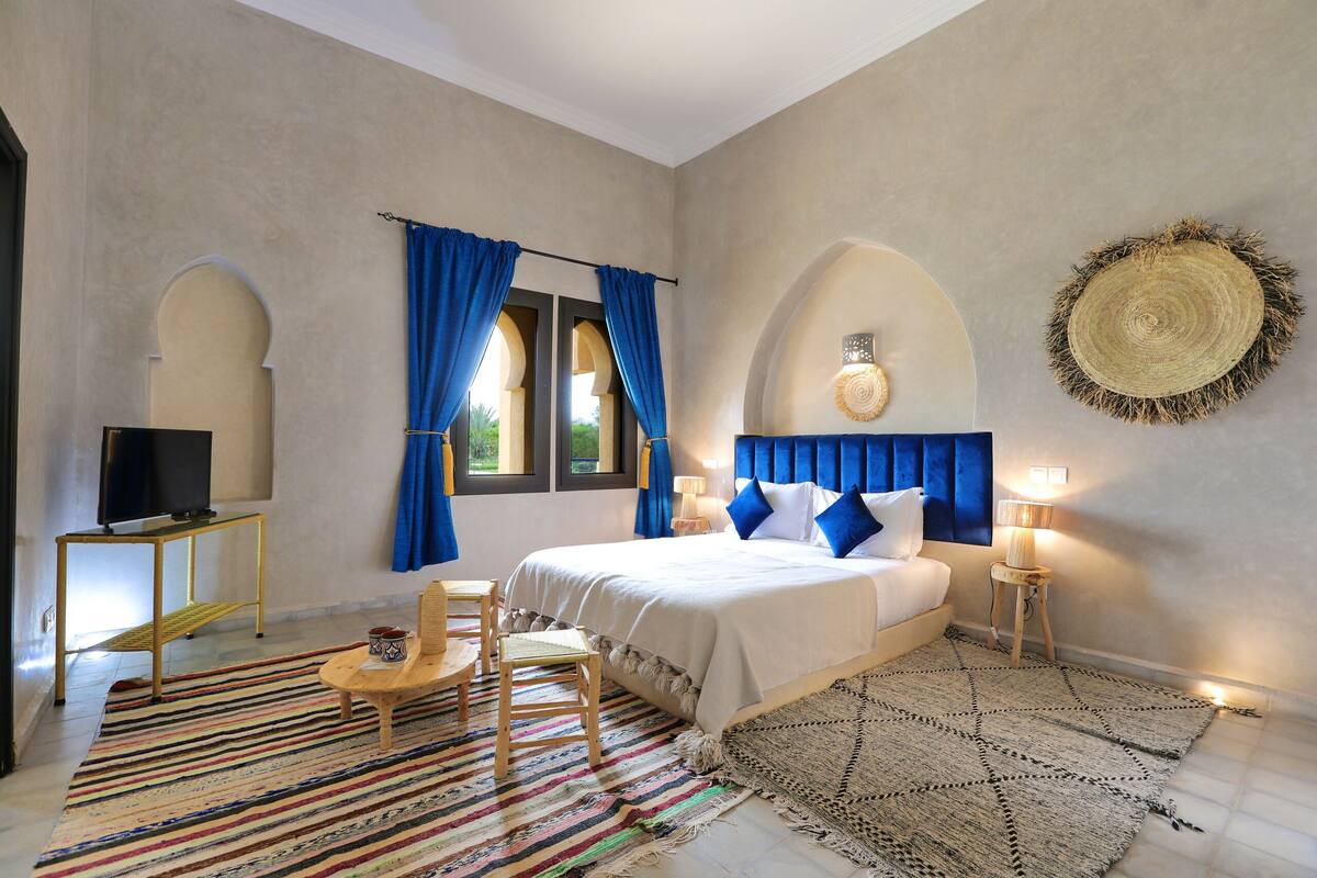 Quarto com cama, televisão, cadeira, mesinha e um objeto decorativo na parede.