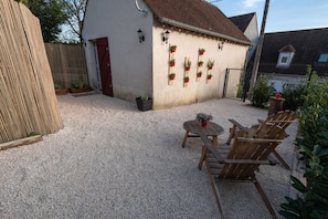 Une terrasse en gravier avec des chaises et une table en bois, un bâtiment blanc avec une porte rouge, et des plantes en pot disposées le long du mur.