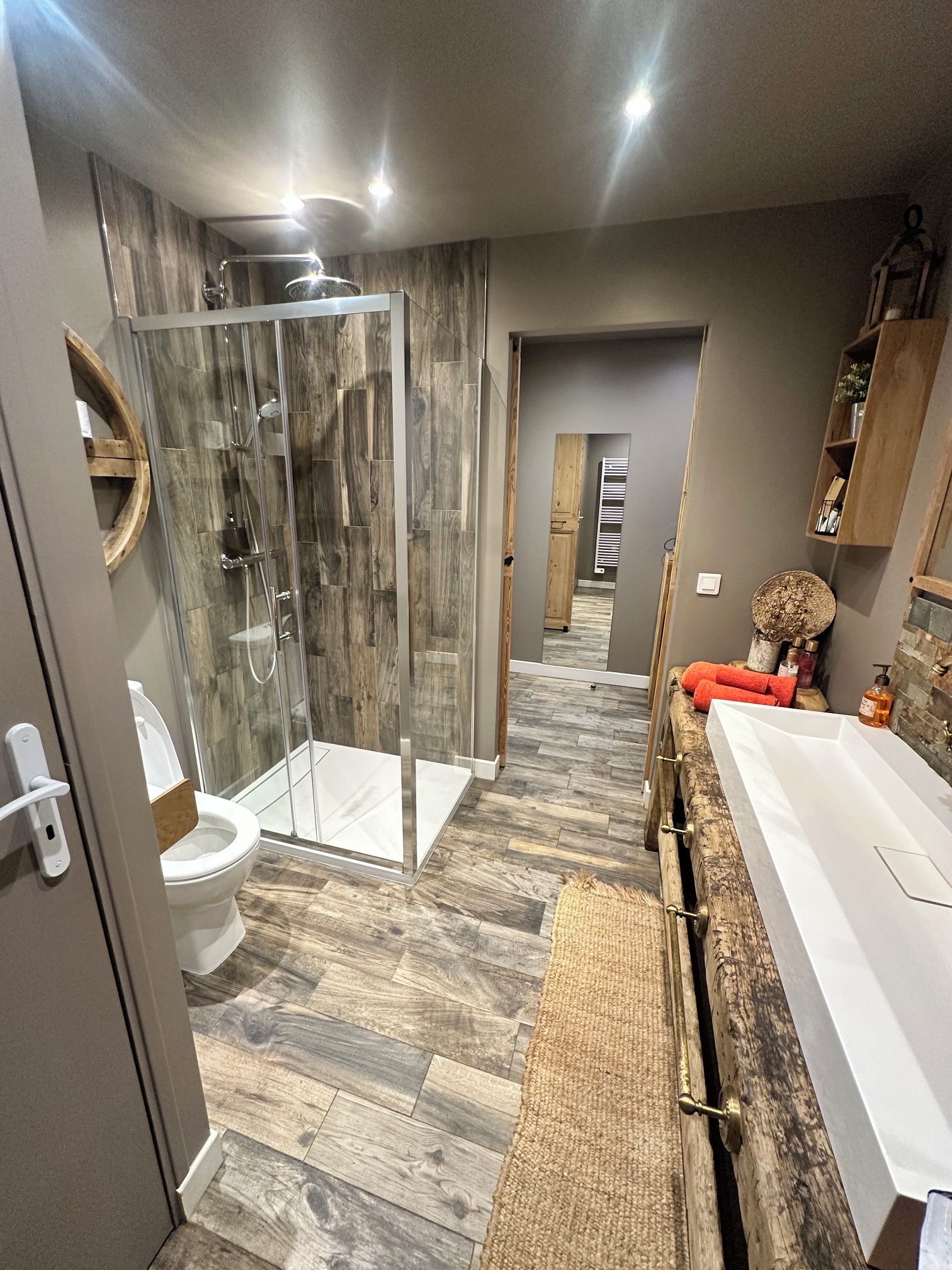 A modern bathroom with a glass shower enclosure, a white toilet, a wooden vanity with a large mirror, and a bathtub.