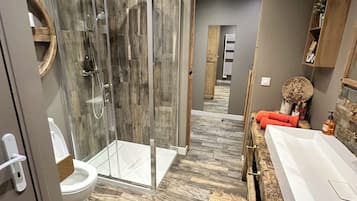 A modern bathroom with a glass shower enclosure, a white toilet, a wooden vanity with a large mirror, and a bathtub.