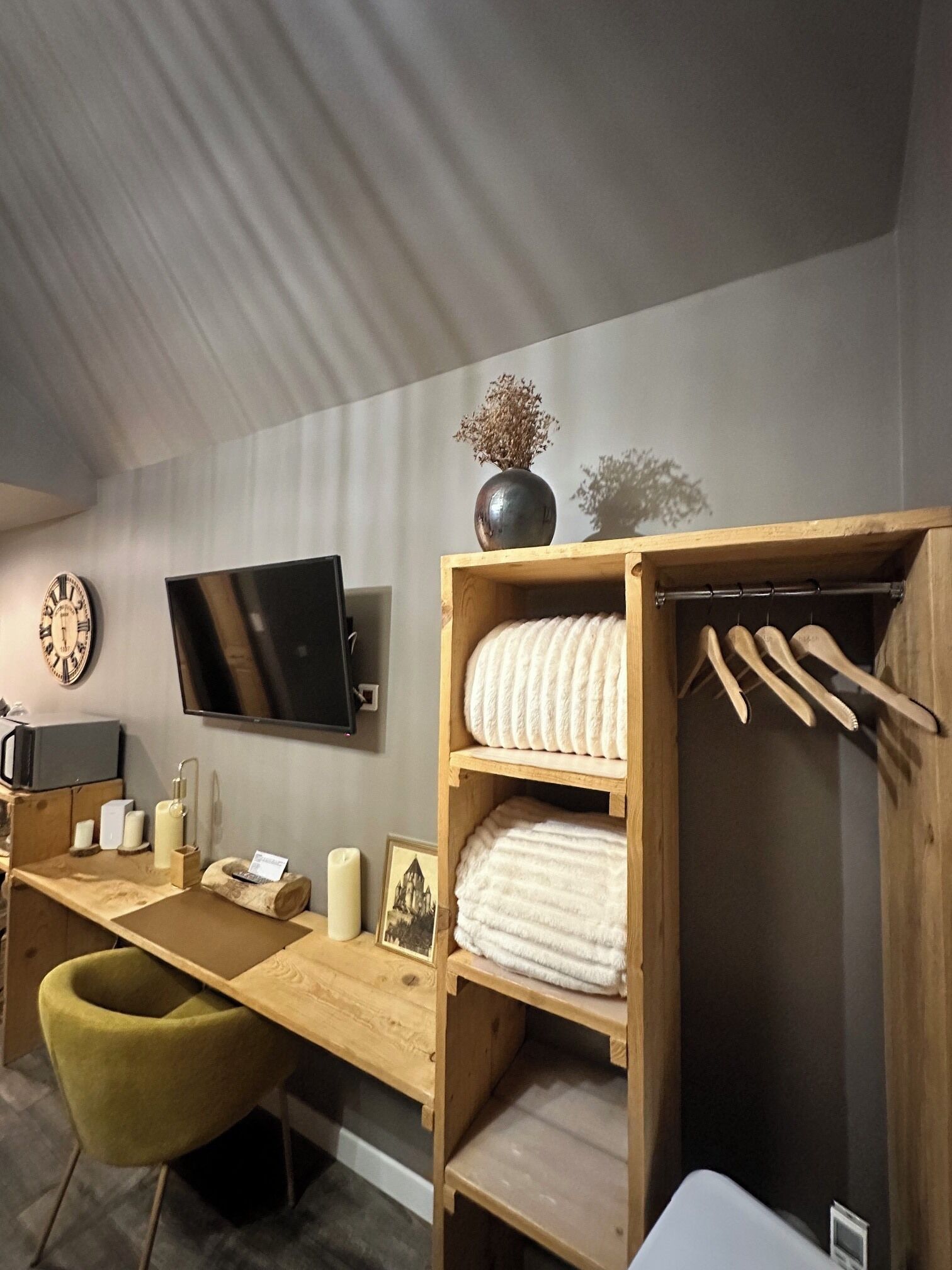 Een kamer met een houten bureau, een aan de muur bevestigde televisie, een houten kledingkast met hangende kleding en een stoel.