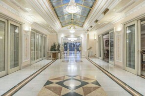 Un couloir d’hôtel avec un sol à motifs géométriques, un plafond en verre et des portes d’entrée disposées de manière symétrique de part et d’autre.
