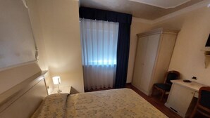 Une chambre d’hôtel avec un lit, un bureau, une chaise, une armoire et une fenêtre avec des rideaux.