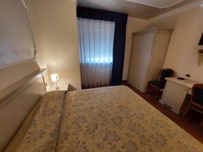 Une chambre d’hôtel avec un lit, un bureau, une chaise, une armoire et une fenêtre avec des rideaux.