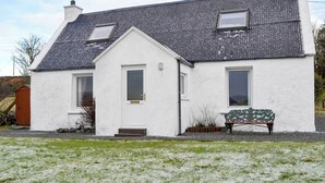 Een wit huis met een grijs dak, twee ramen, een deur en een bankje in de voortuin.