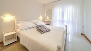 A neatly arranged bedroom with a bed, bedside tables, and a large window with sheer curtains. - Apartments Loncar (Krk)